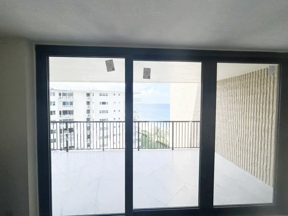 A view from inside through large glass sliding doors onto a balcony overlooking a high-rise building and the ocean.