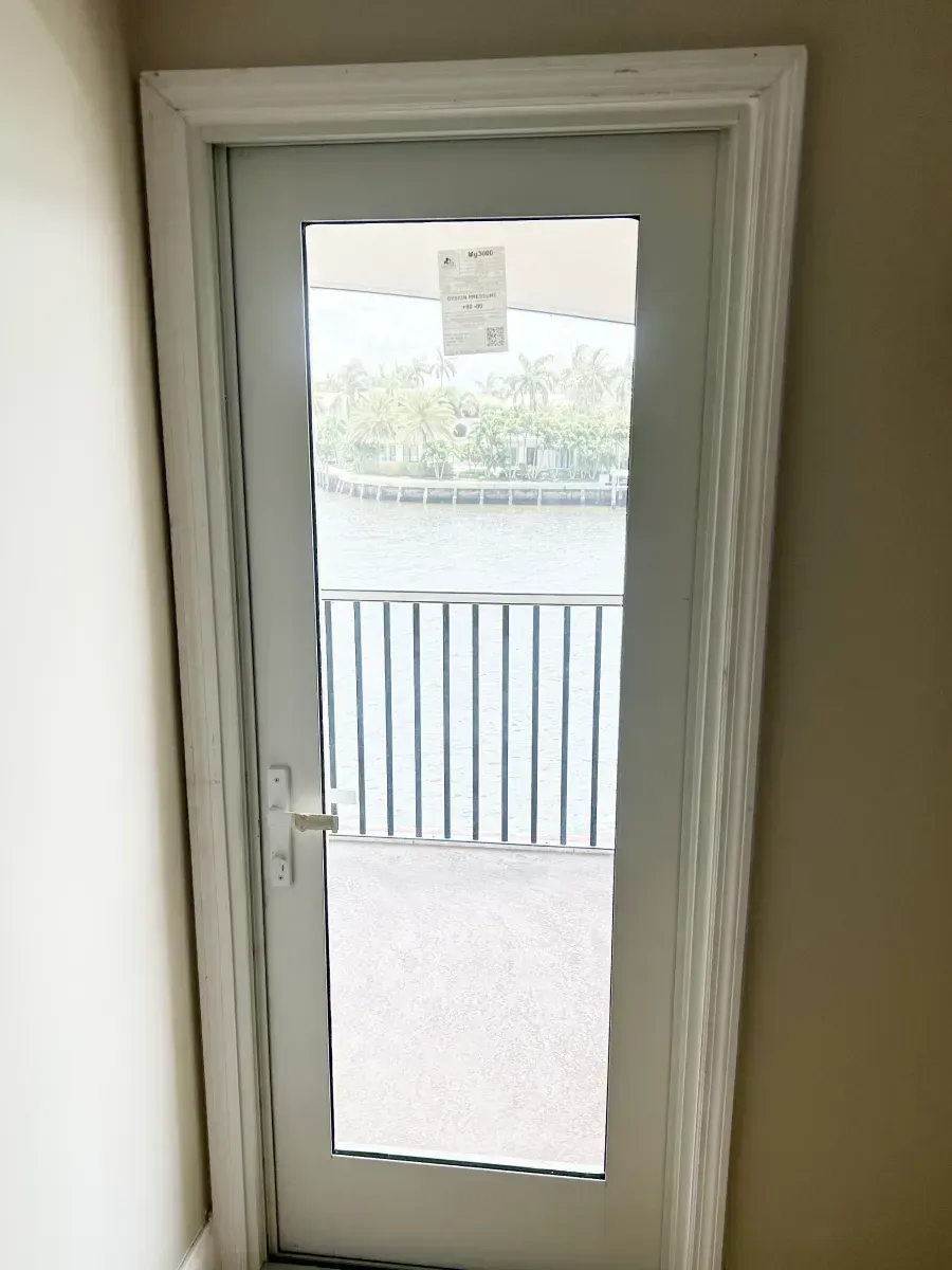 A white-framed glass door leading to a balcony with a view of a body of water and palm trees.