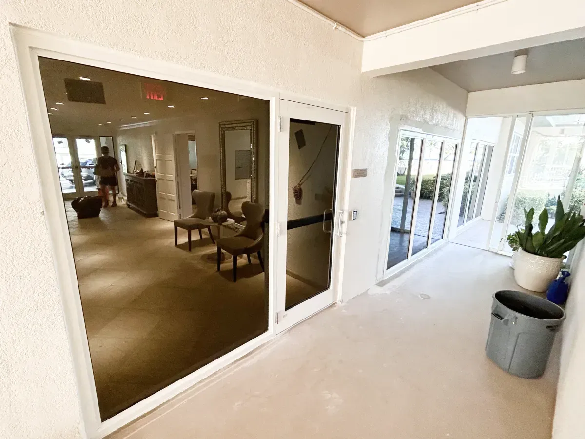 A bright hallway with a glass-fronted room containing chairs, a large mirror, and a person walking in the distance.
