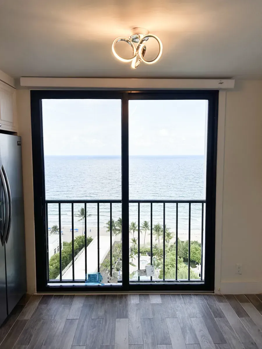 A sliding glass door with a black frame looking out onto a beach, ocean, and palm trees, with a modern ceiling light above.