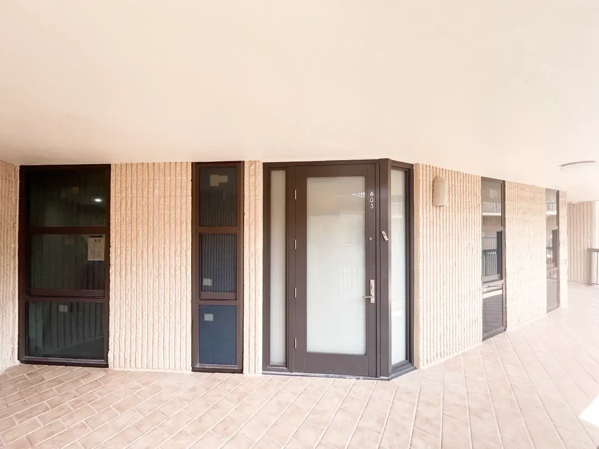 Building entrance with dark-framed windows and a glass door, set in a beige, textured wall on a patterned tiled walkway.
