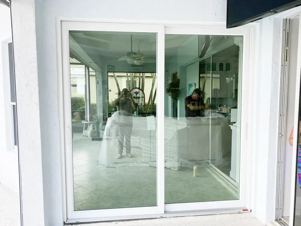 A white sliding glass door with interior reflections showing two people standing in a room with light-colored walls.
