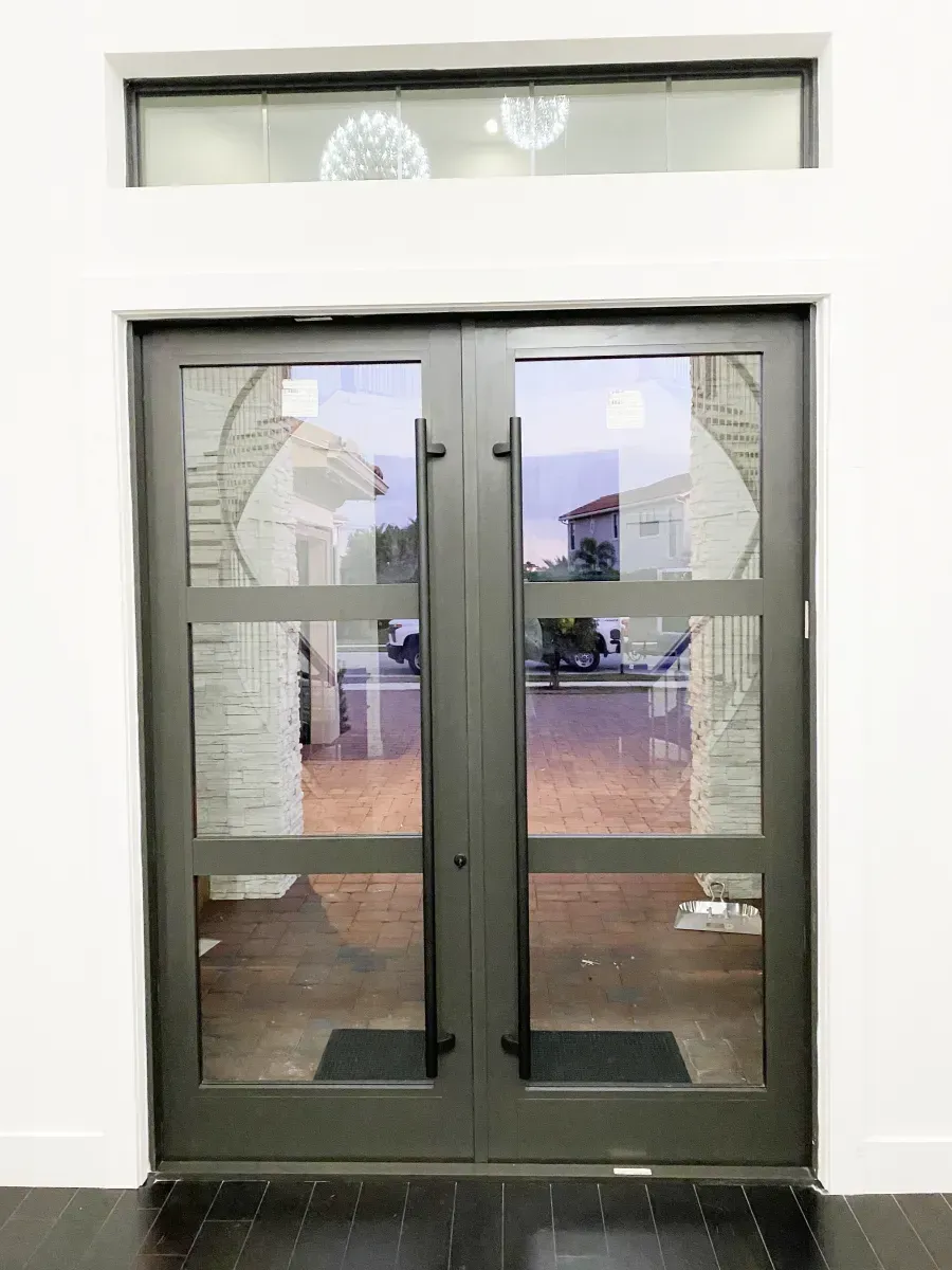 Double dark gray metal doors with glass panels and a horizontal transom window above, set against a white wall.