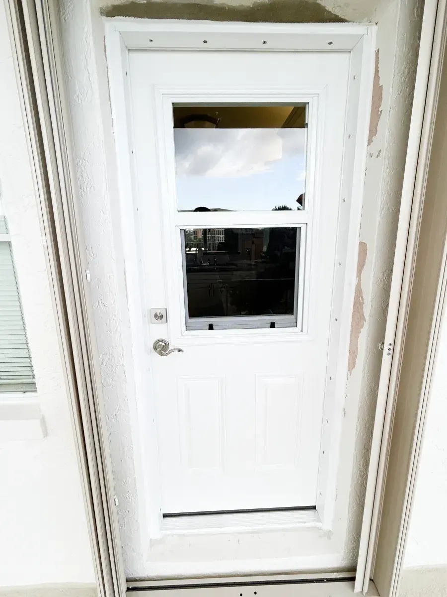 White exterior door with a rectangular window, silver handle, and deadbolt, set in a light-colored building frame.