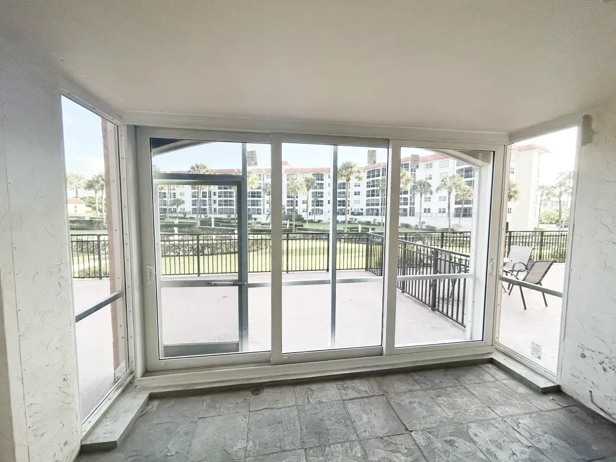 A bright, screened-in balcony with tile floors looking out toward a large white building and palm trees.