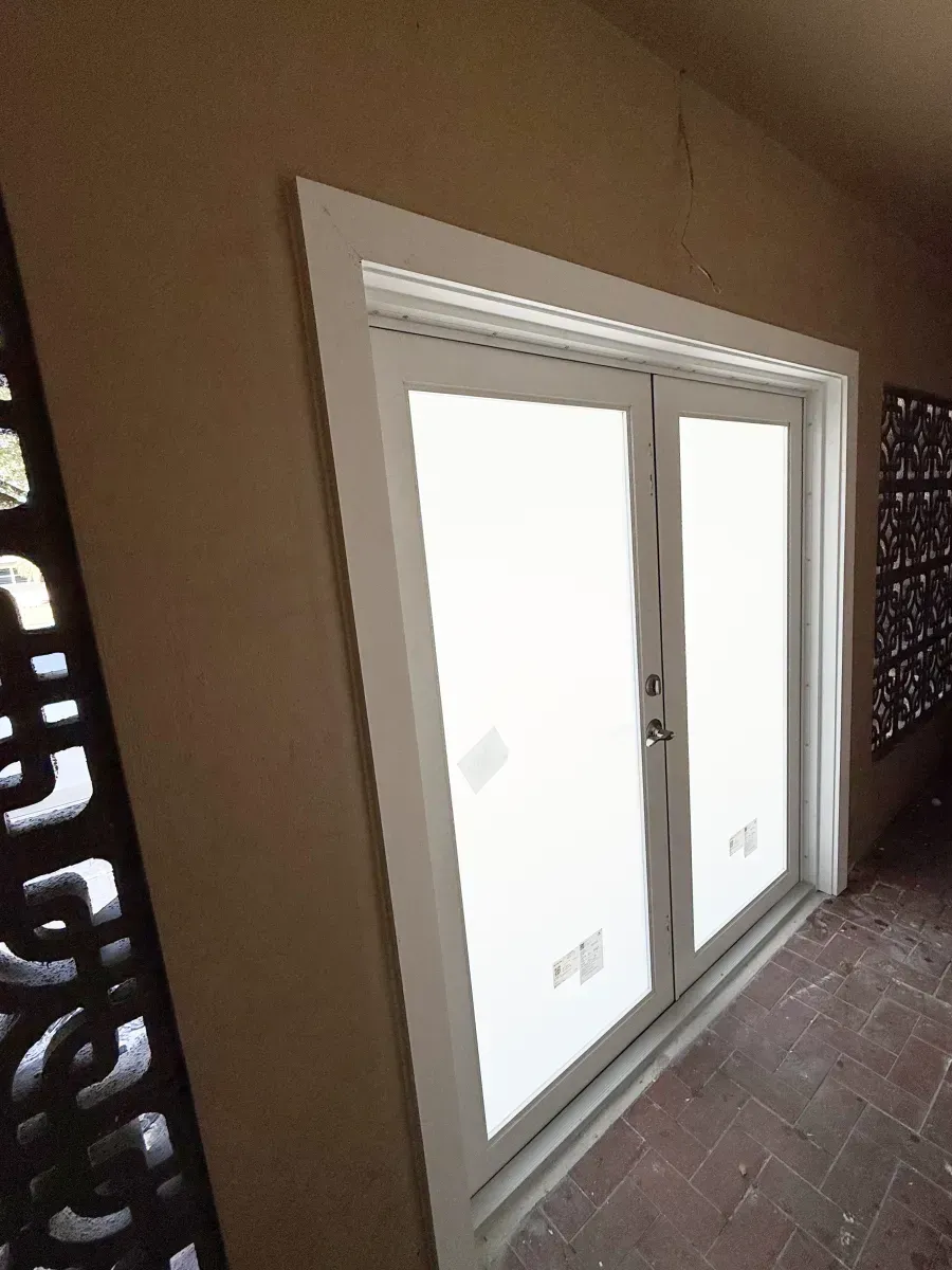White double doors with glass panels set in a tan wall next to decorative dark metal screens on a tiled patio.