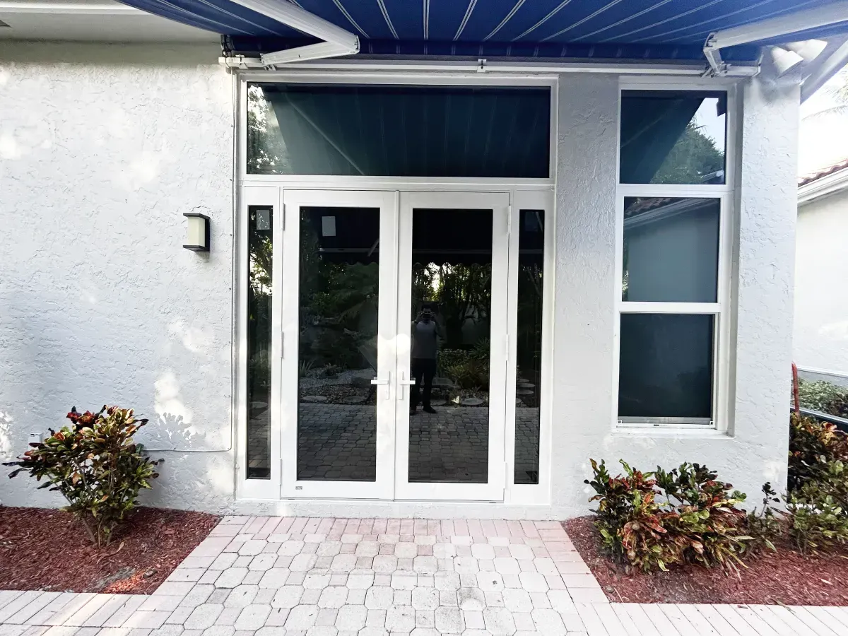 A white double door entry with glass panels, a large transom window, and a side window on a light-colored stucco building.