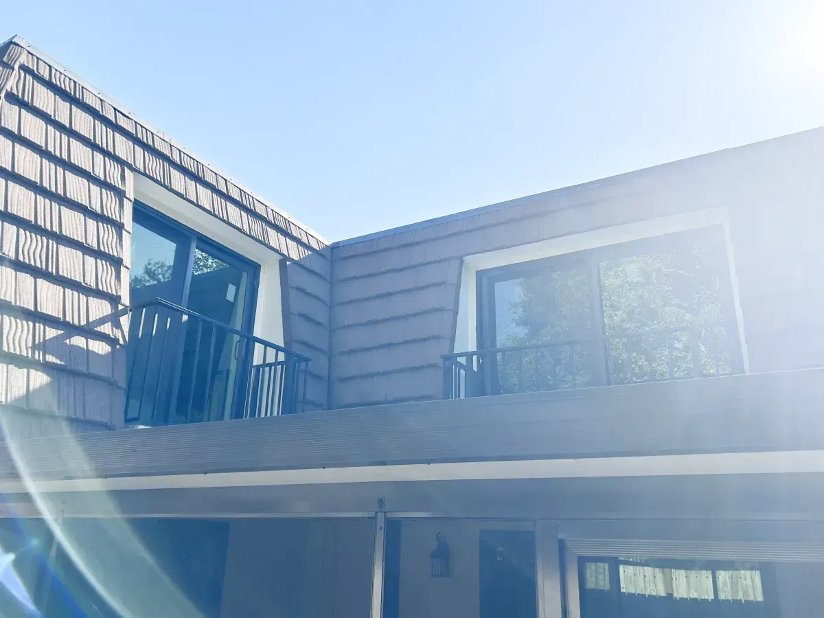A low-angle view of a two-story residential building with brown wood-shake siding and two balconies under a clear sky.