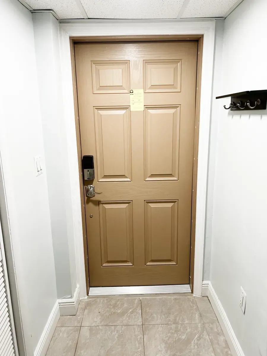 A tan, four-panel interior door with a digital keypad lock, centered in a light gray hallway with a wall-mounted coat rack.