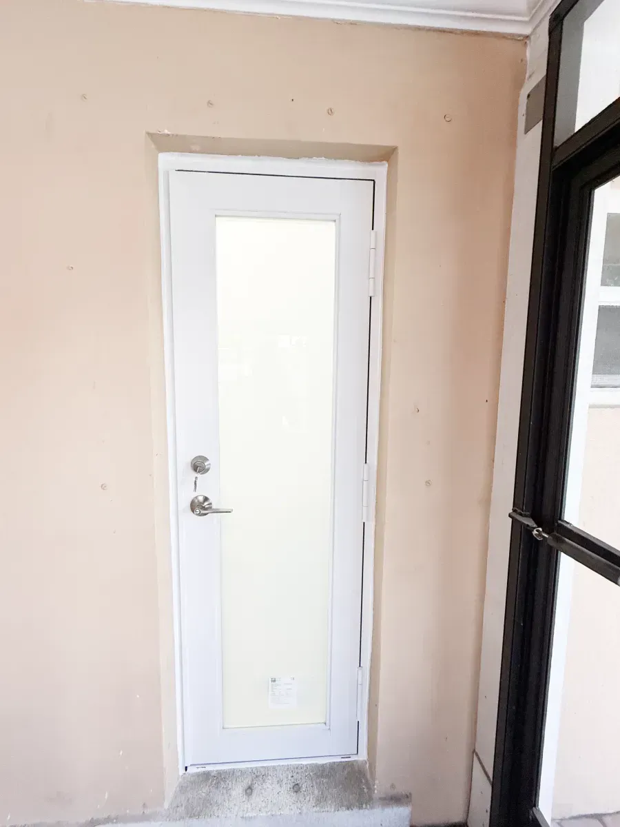 A white, narrow door with a vertical glass panel set into a plain, light-beige interior wall.