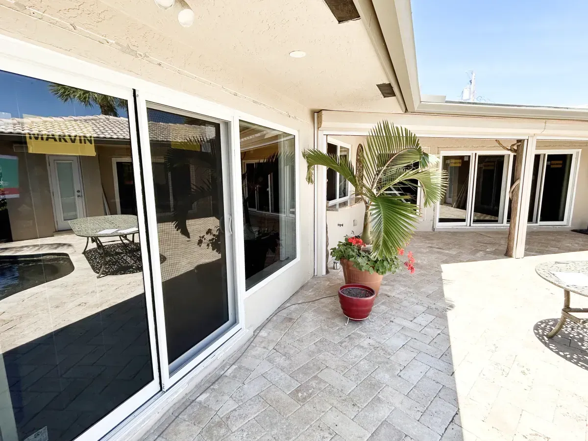 A patio area with light-colored pavers, sliding glass doors, and a potted palm plant under a roof overhang.