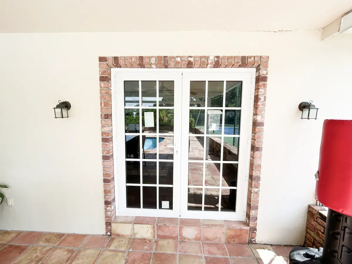 White French doors framed by brick, centered on a light-colored wall with two wall sconces and terracotta tile flooring.