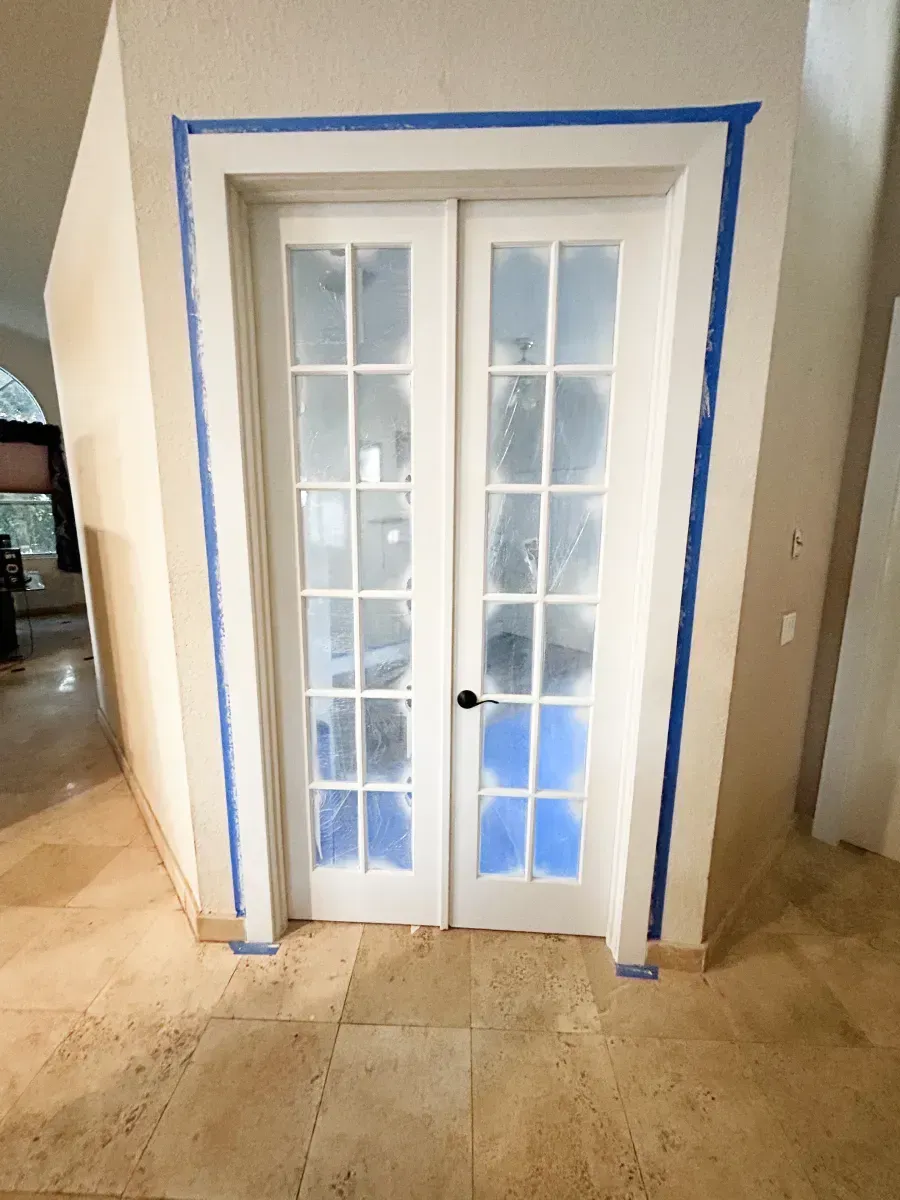Double French doors with white frames and blue painter's tape surrounding the trim, set against a beige wall and tile floor.