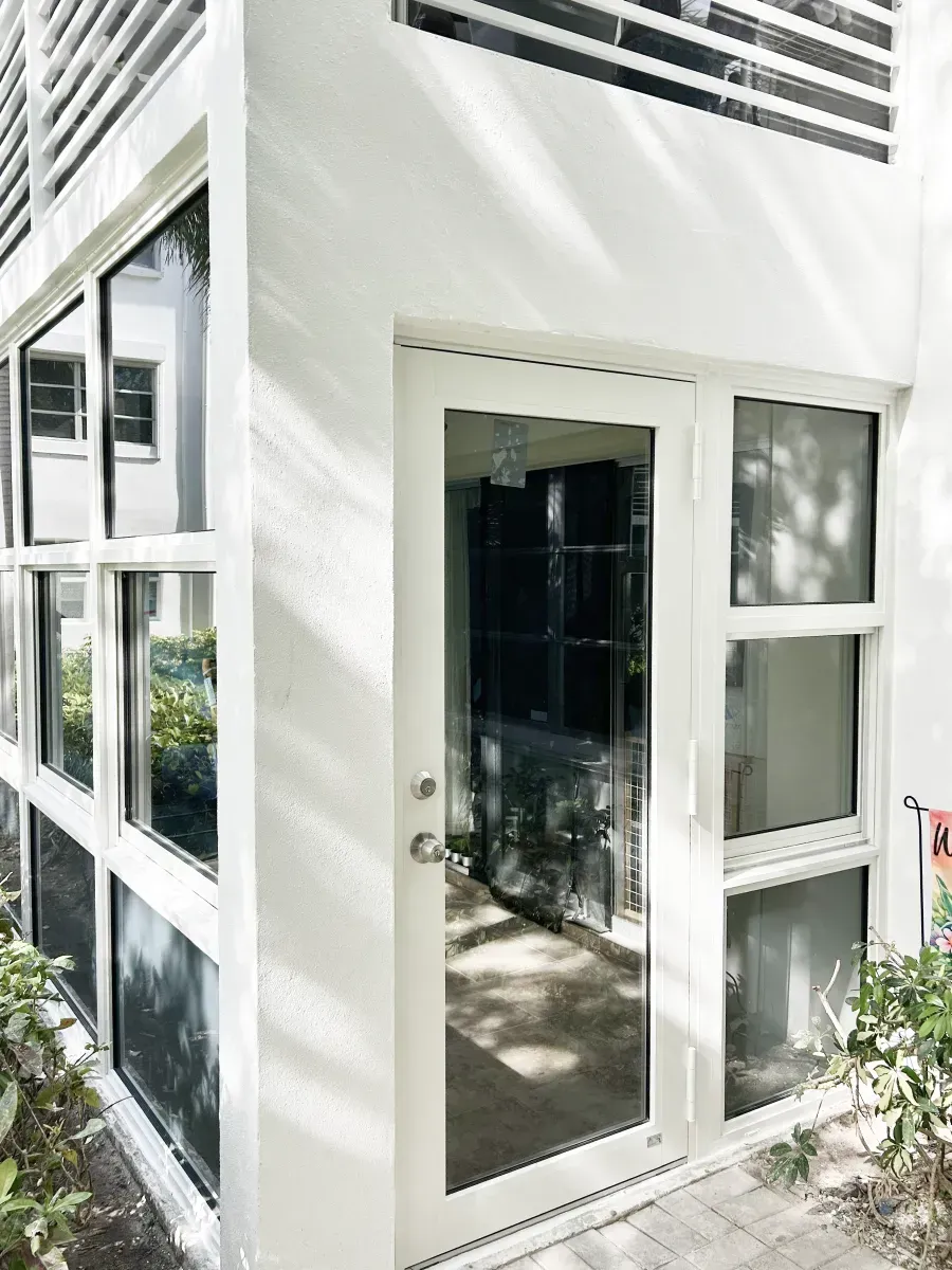 A white building corner features a glass-paned door, large windows, and horizontal slats on the upper level.