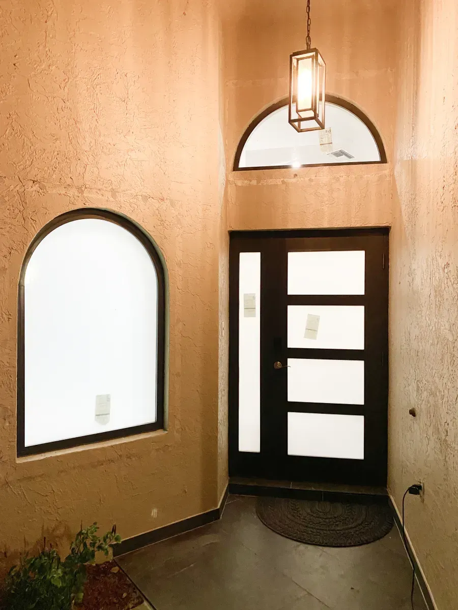 A tan stucco entryway with a dark-framed modern front door, an arched window, and a lantern-style pendant light.