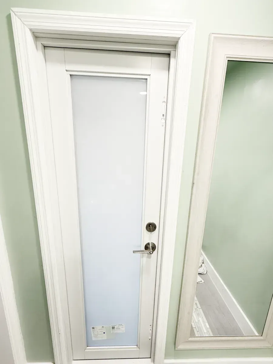 A white door with a frosted glass panel set in a light green wall next to a large, matching white-framed floor mirror.