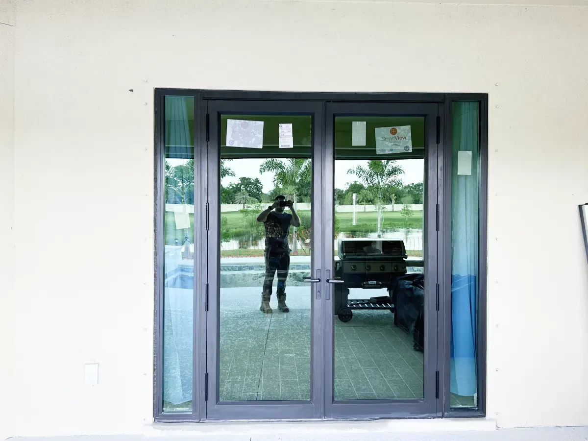 A double glass door with dark gray frames set in a plain white wall, reflecting a person taking a photo and an outdoor area.