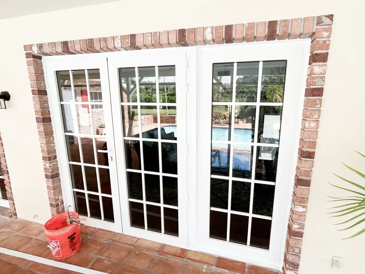 White French doors with multi-pane windows set in a brick frame on a tiled patio, with a red bucket nearby.