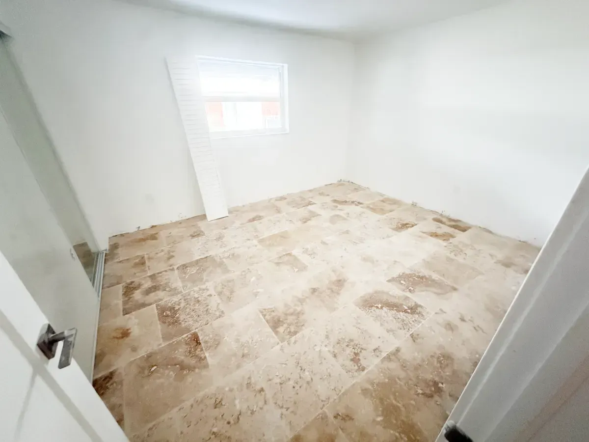 An empty room with white walls and light brown, textured stone tile flooring, viewed through an open doorway.