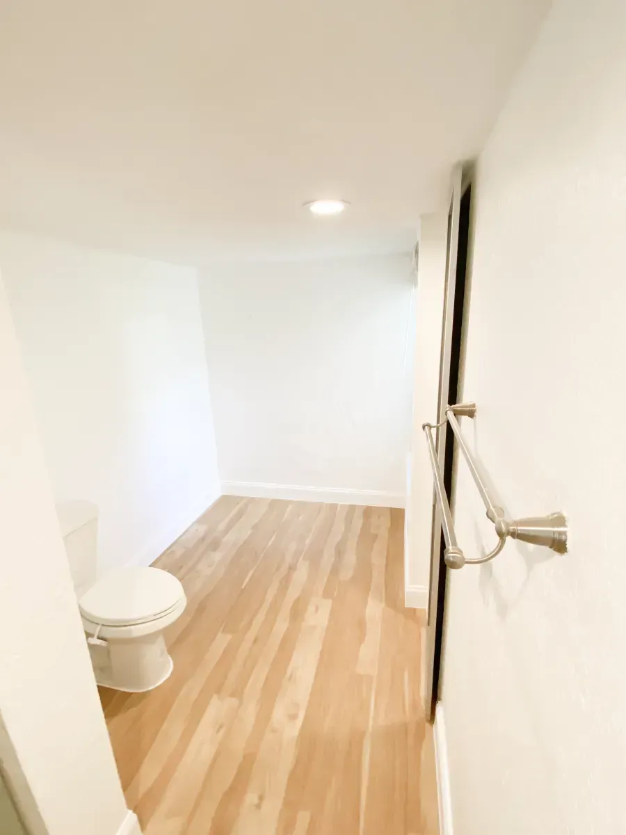 A bathroom with light wood-look flooring, a white toilet, and a brushed nickel towel bar on a white wall.