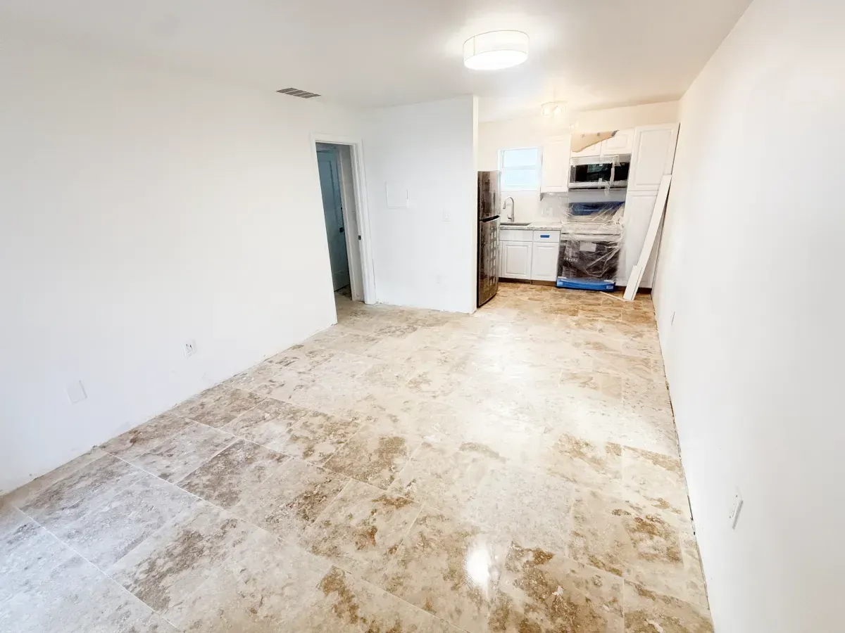 A bright, empty room with large, mottled beige tile floors and white walls, leading into a kitchen with stainless steel.