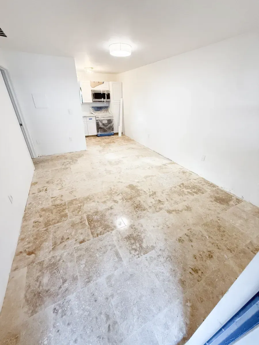 An empty room with white walls and light-colored stone tile flooring, leading to a small kitchen area in the background.