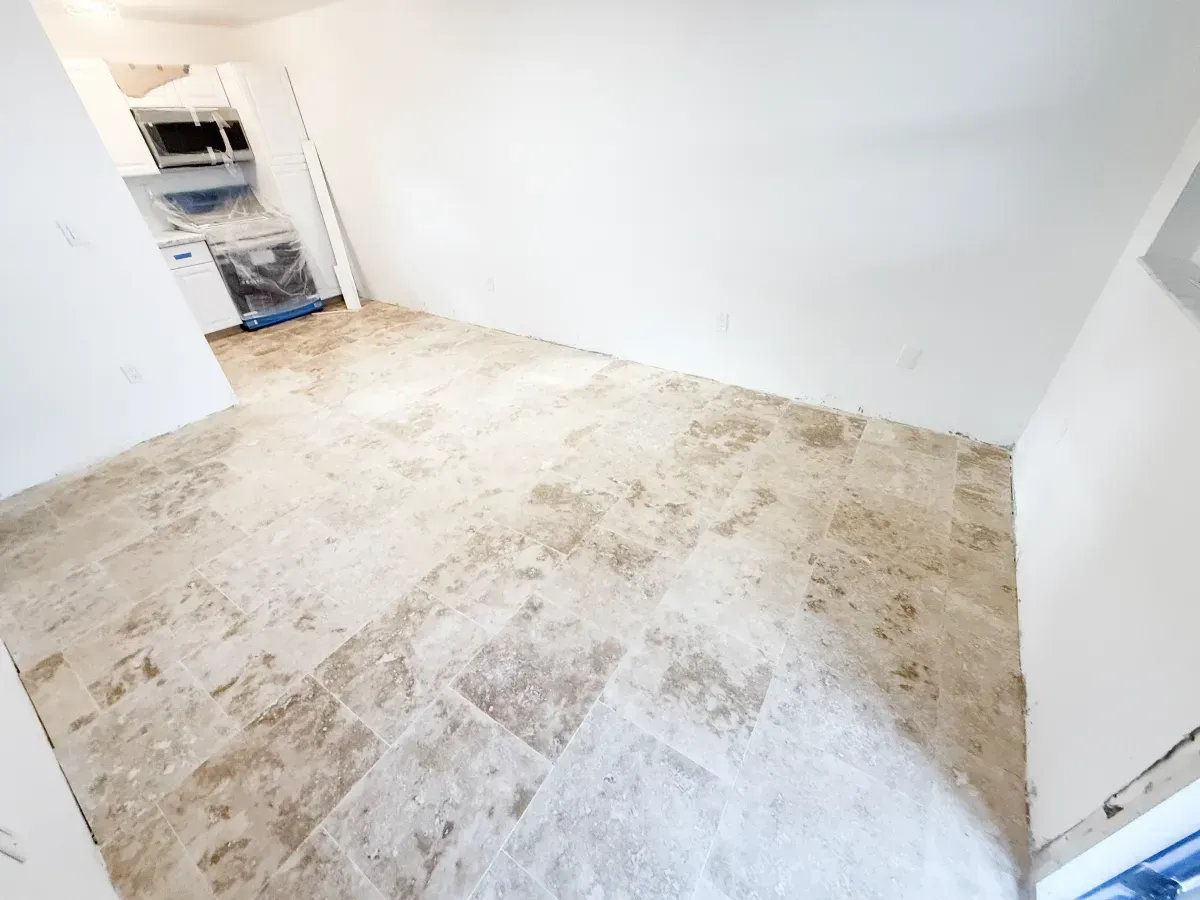 An interior view of a room under renovation with light brown travertine tile flooring and bare white walls.