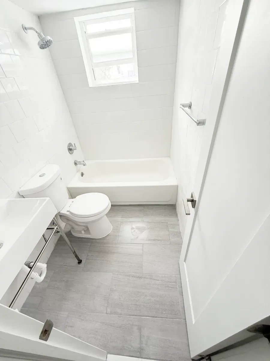 A small, white bathroom featuring a shower-tub combination, pedestal sink, and gray tiled flooring.