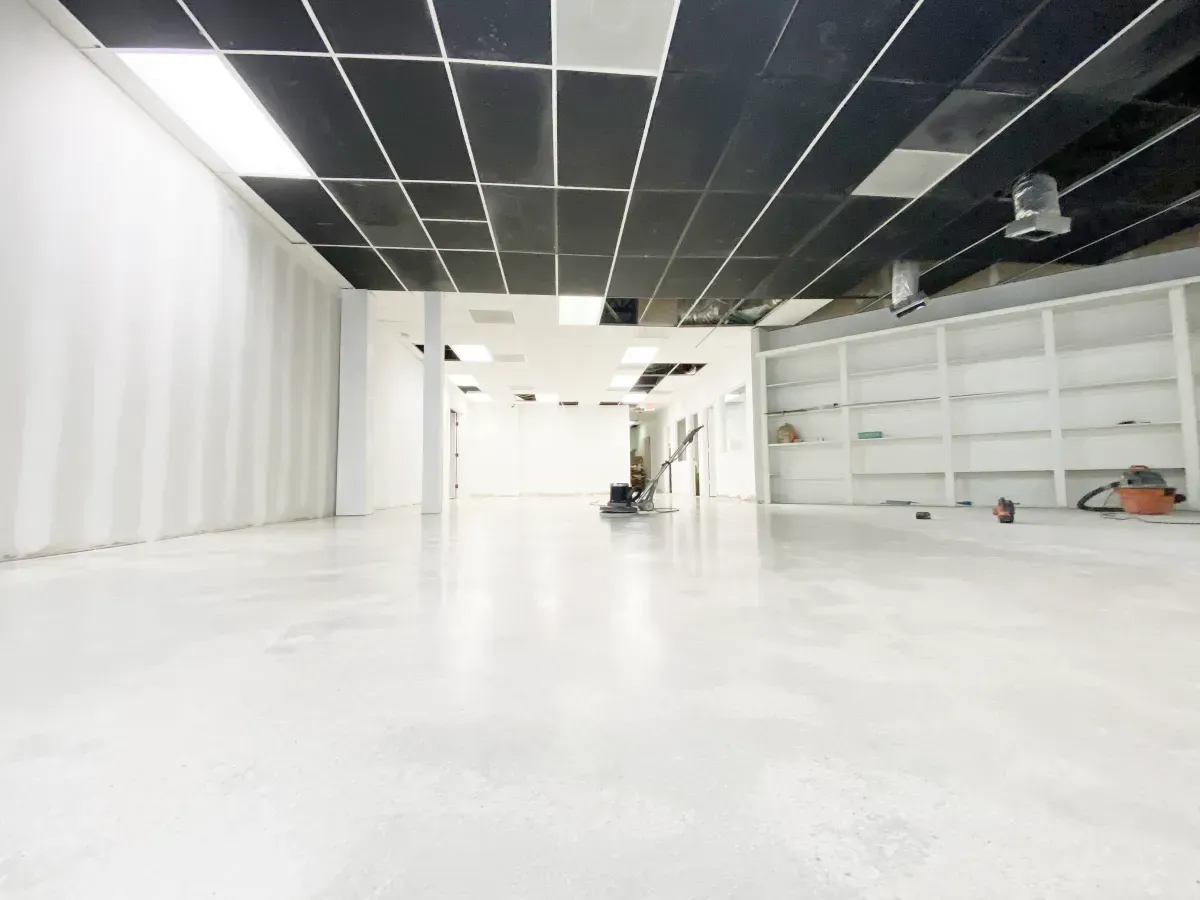 An empty commercial space with a glossy white concrete floor, black drop ceiling, and white shelving against the wall.