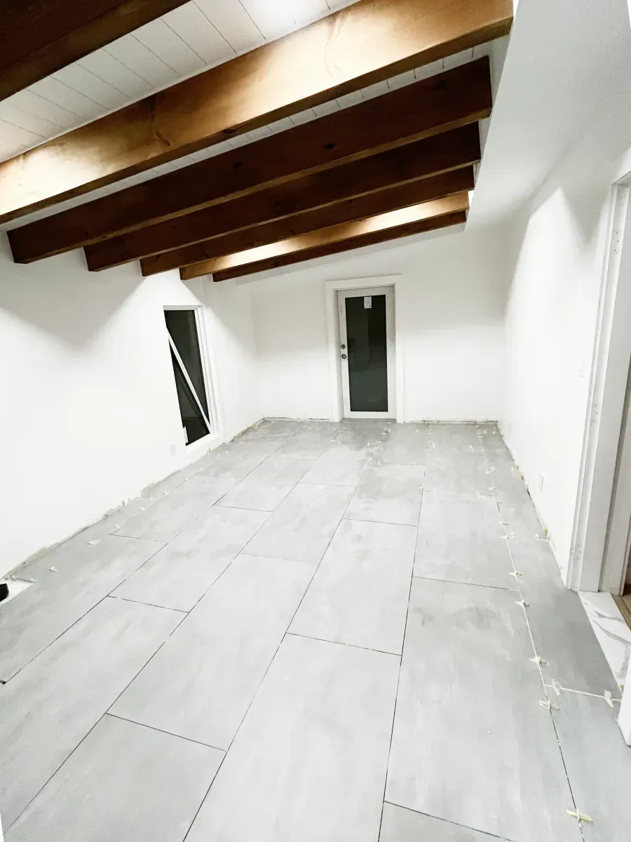 A sunlit, minimalist room with wooden ceiling beams, white walls, and large grey floor tiles leading to a back doorway.