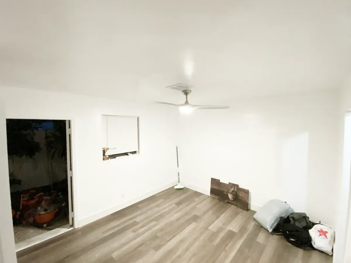 An empty room with white walls, a ceiling fan, wood-look flooring, a doorway, a small window, and bags on the floor.