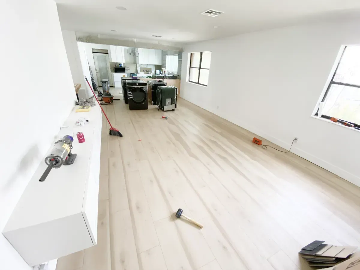 A wide view of a bright, white, unfurnished room under renovation with light wood-tone flooring, tools, and a distant kitchen.
