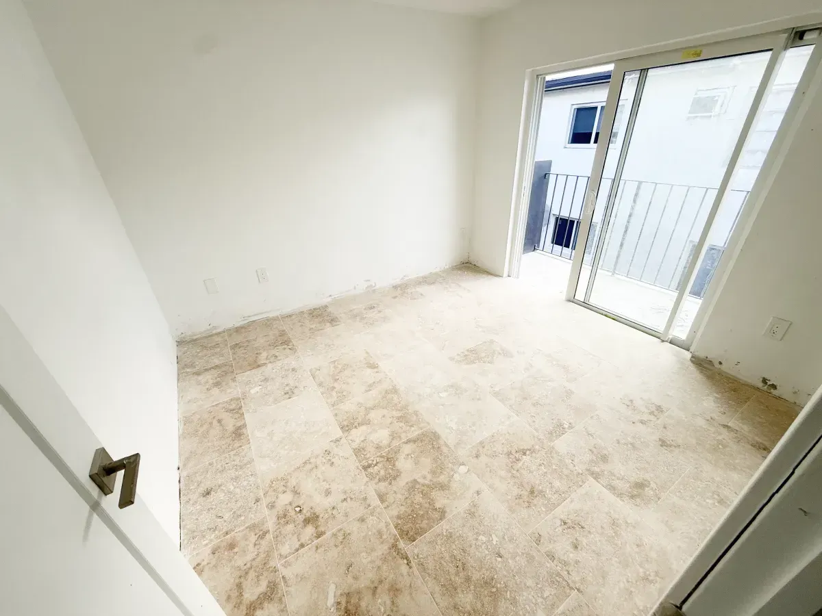 An empty room with light-colored tile flooring and white walls, featuring a sliding glass door leading to a balcony.