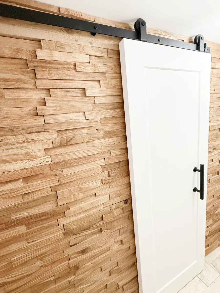 A white sliding barn door with a black handle mounted on a wall with textured, natural wood paneling.