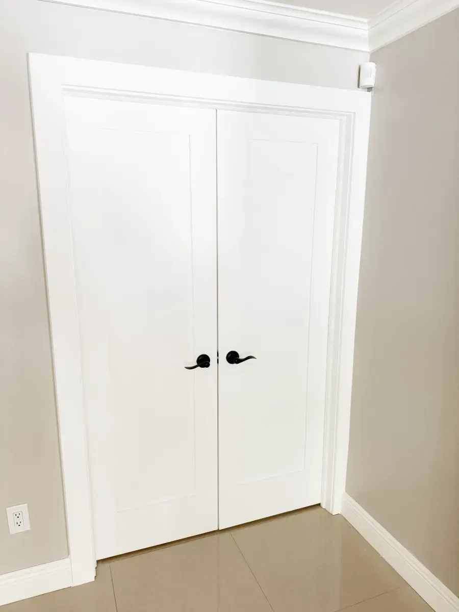 White double doors with black handles in a hallway with neutral walls and light-colored tile flooring.