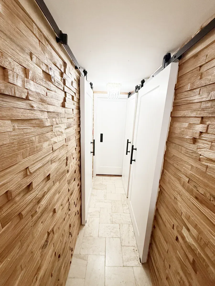 A narrow hallway features textured wooden slat walls, light tile floors, and several white sliding barn doors.