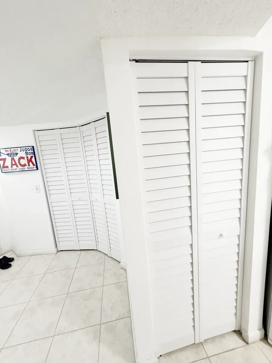 White bi-fold louvered closet doors stand in a room with light-colored tile floors and a wall sign that reads ZACK.