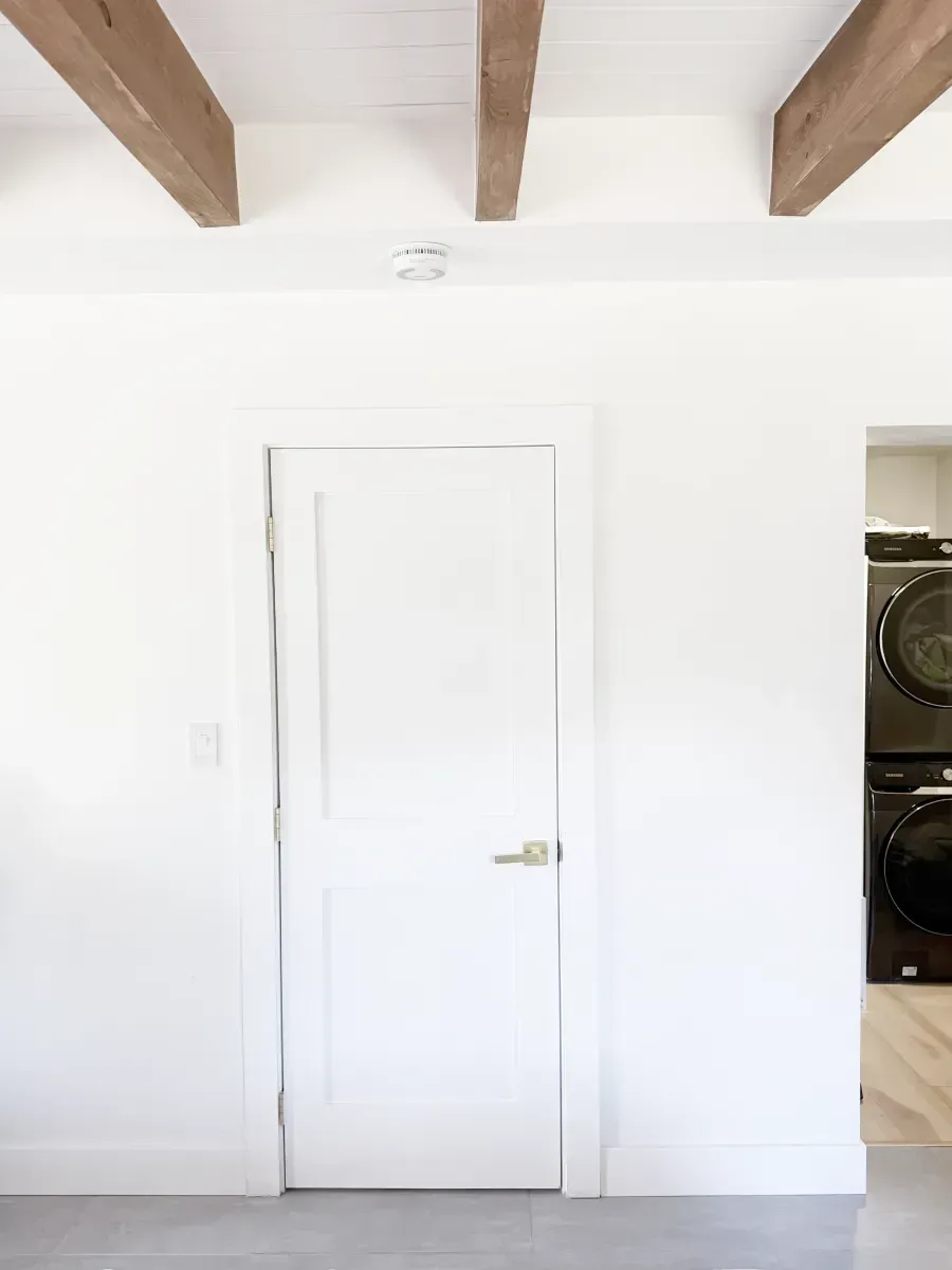 A white door stands centered against a white wall with wooden ceiling beams above, with a laundry area visible to the right.