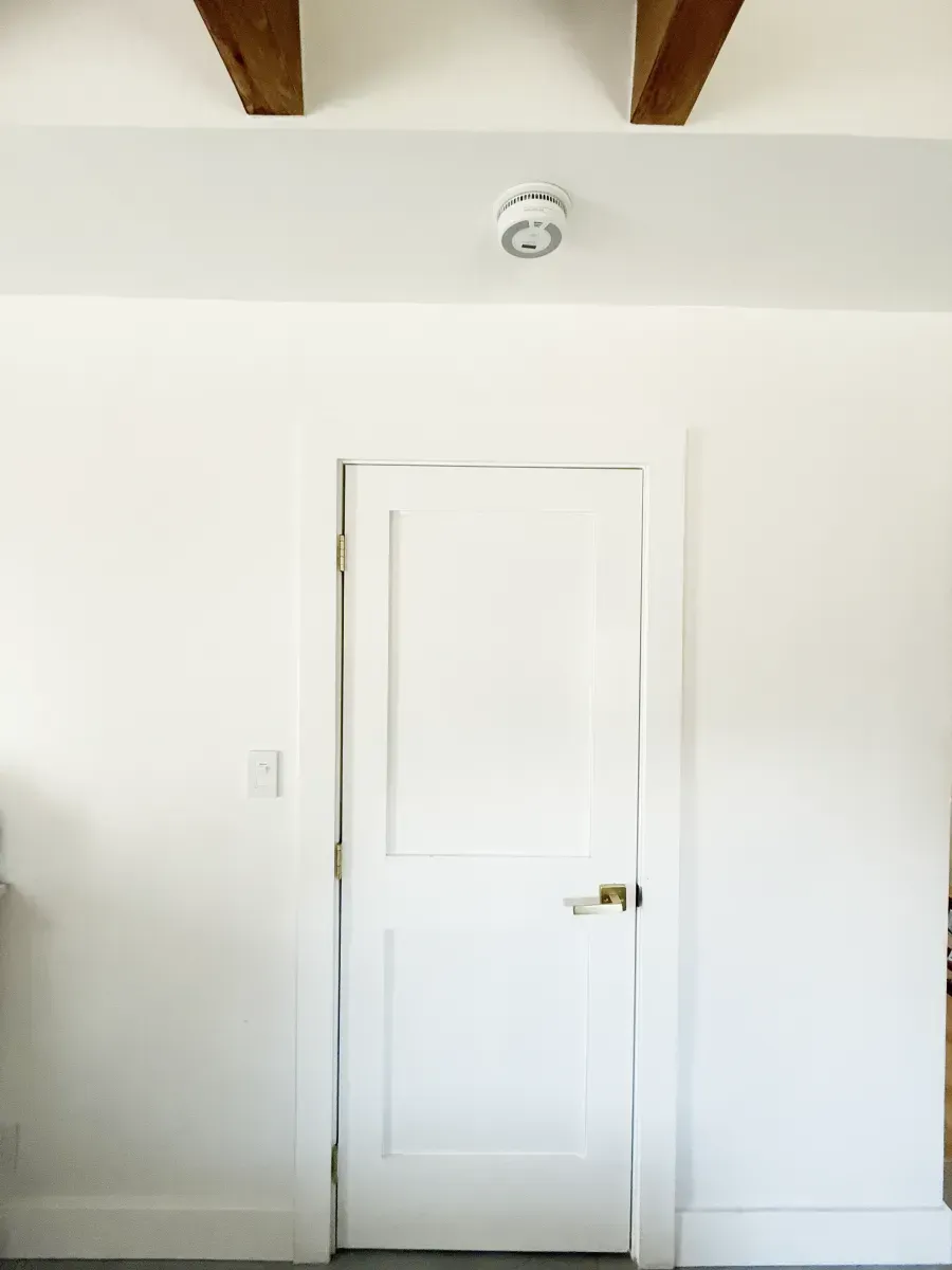 A white door set in a white wall with two wooden ceiling beams and a smoke detector mounted above the door frame.