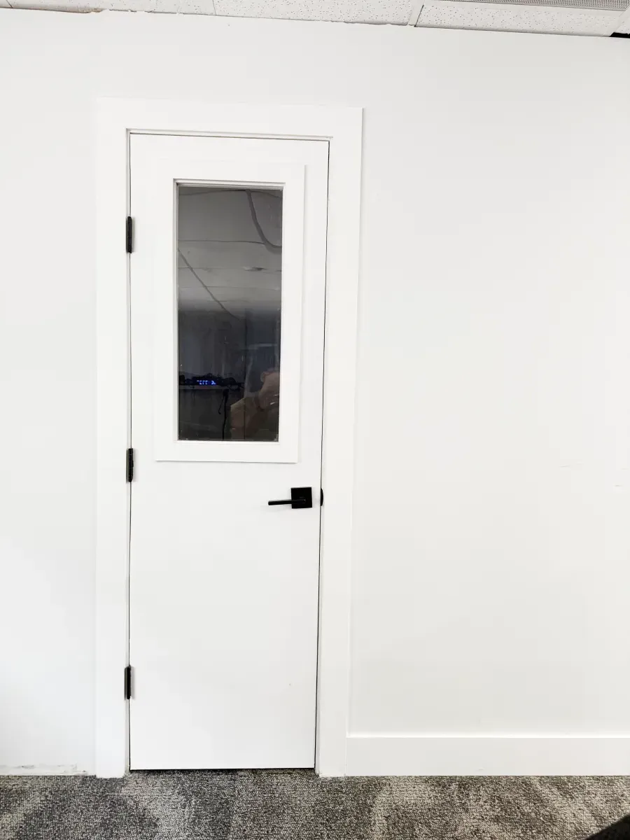 A white, single-panel door with a rectangular glass window and black handle installed in a white wall above gray carpet.