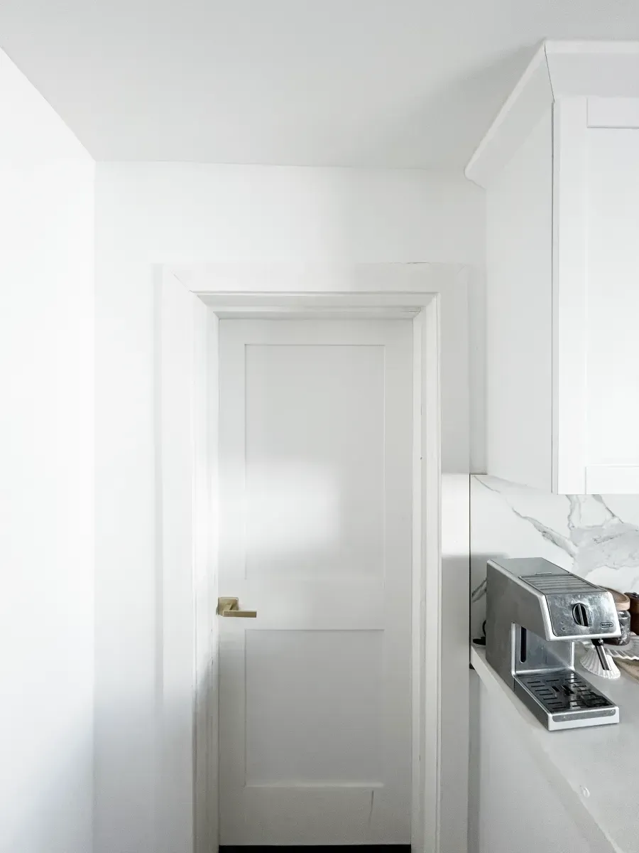 A white, two-panel door stands in a bright, minimalist room next to a metallic coffee machine on a white counter.