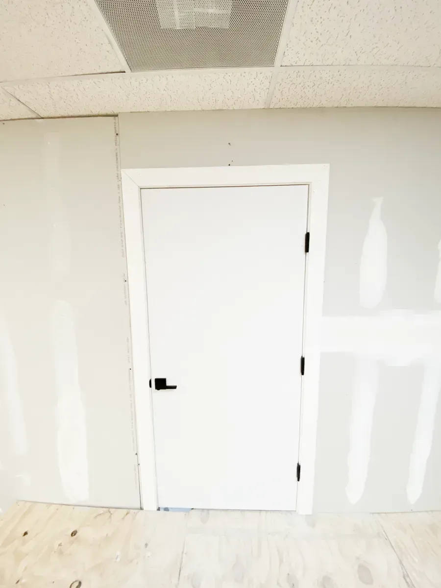 A white door with a black handle set in an unfinished room with drywall walls and a plywood floor.