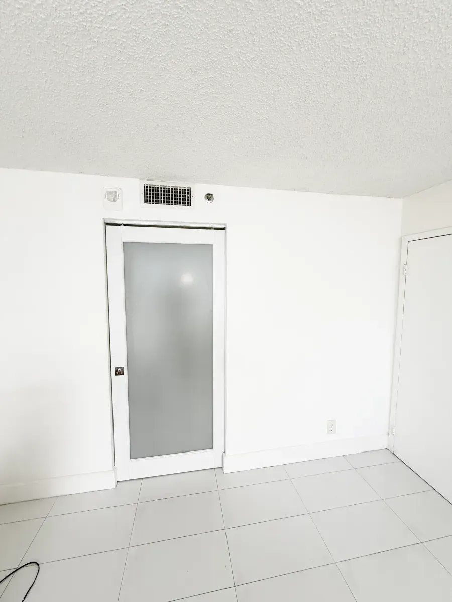 A room with white tile floors, a white wall, and a central frosted glass door beneath a small HVAC vent.