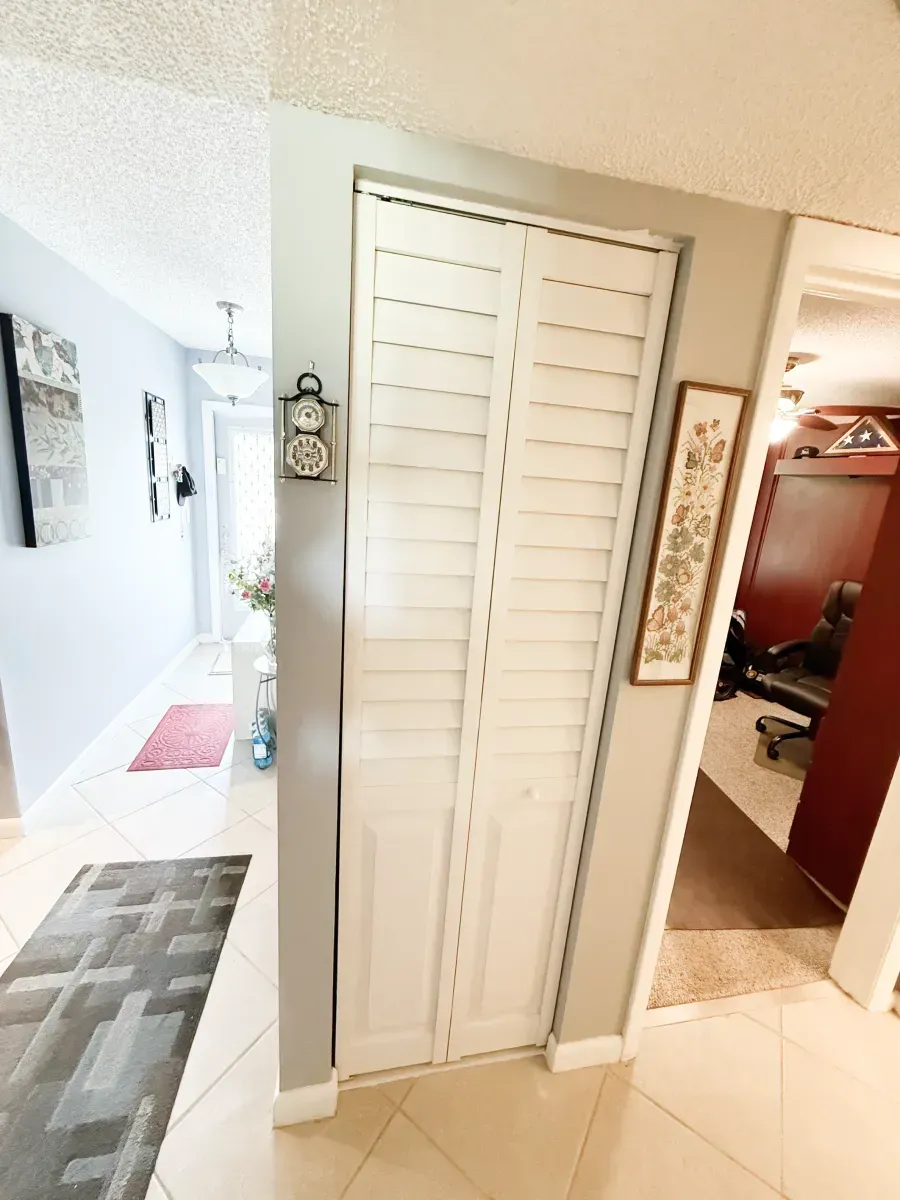 A white louvered bifold door stands on a wall between a tiled hallway and a room with dark wood furniture and carpeting.
