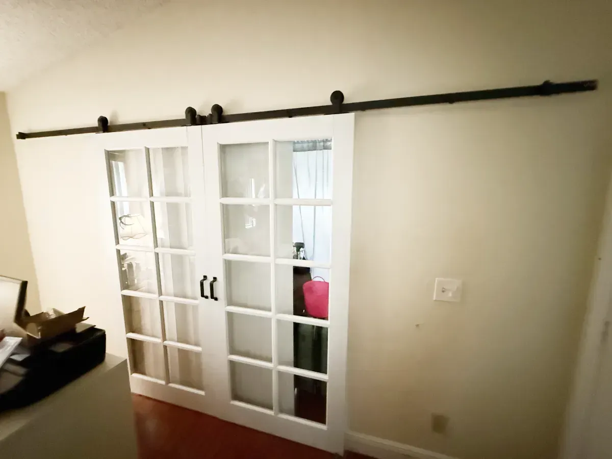 A pair of white, glass-paned French barn doors hanging on a black metal sliding track against a neutral wall.