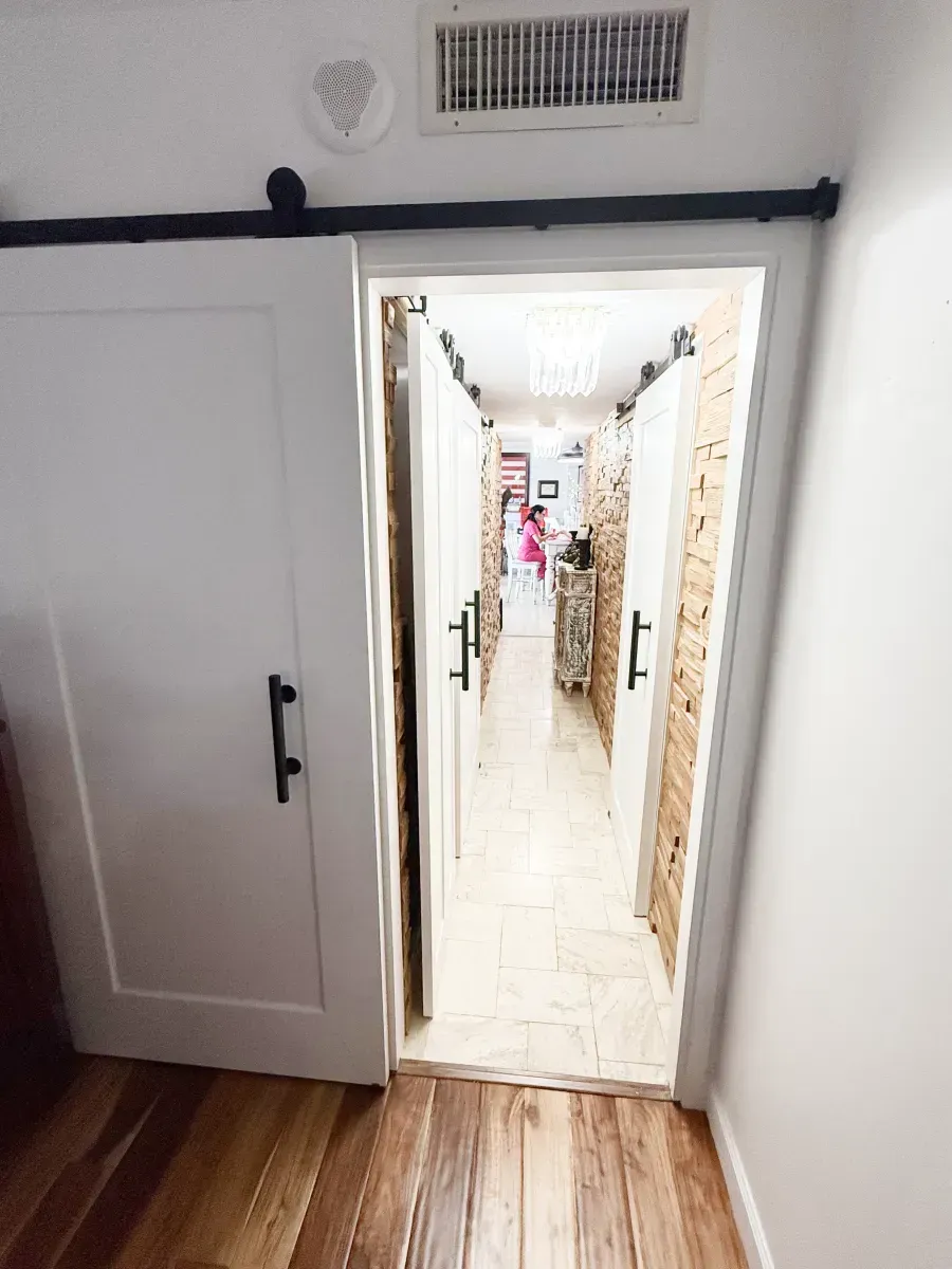 A white barn door opens into a hallway with stone walls, a tile floor, and another barn door in the distance.