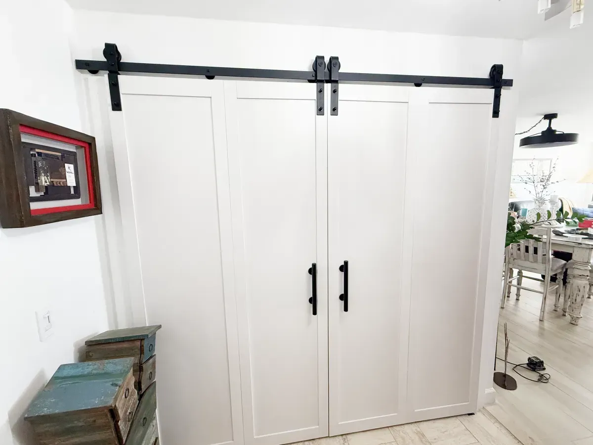 Double white barn doors with black hardware and vertical handles mounted on a black sliding rail in a home interior.