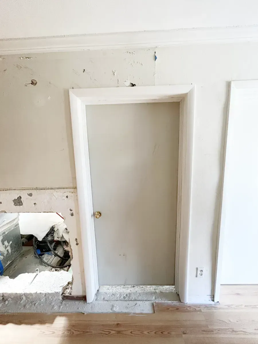 An unfinished interior wall with a door, showing bare drywall, construction debris, and a patch of smooth, painted wall.