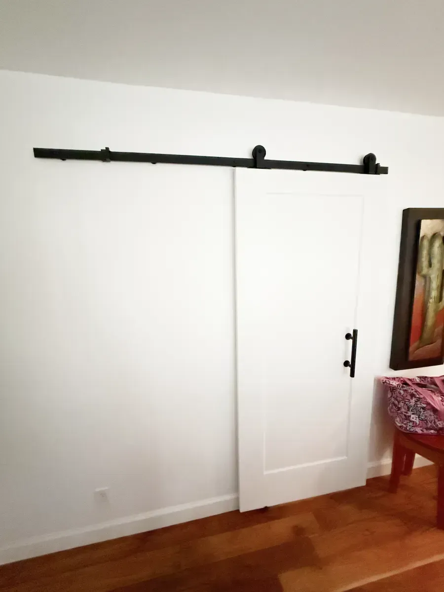 A white interior sliding barn door with black hardware mounted on a white wall above a wooden floor.