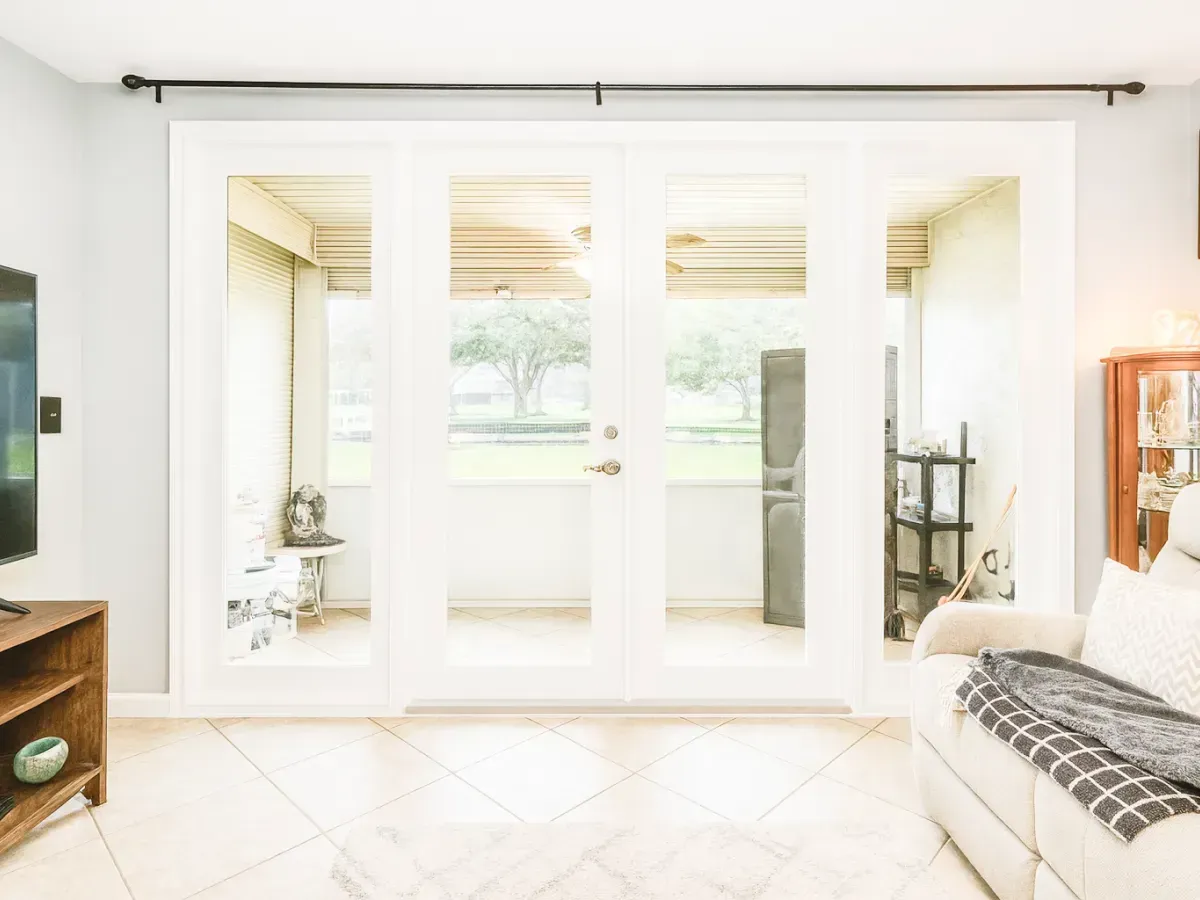A light-filled living room featuring large white-framed glass doors leading to a patio, tile floors, and a sofa.